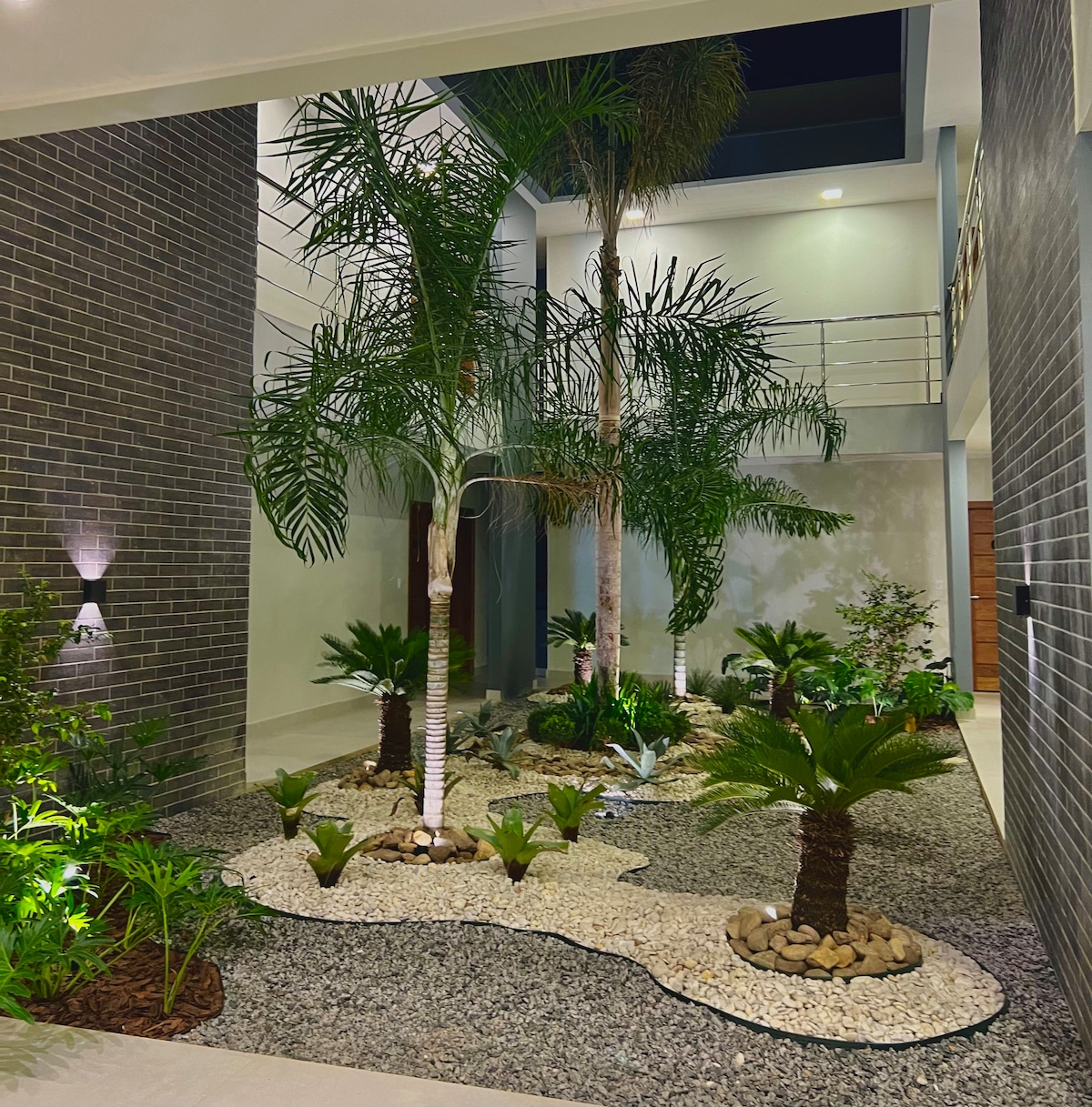 An indoor garden is featured, showcasing tall palm trees and various plants set within a pebble-strewn landscape. Soft lighting highlights the greenery, creating a tranquil and inviting atmosphere. The space is framed by modern brick walls and open staircases above.