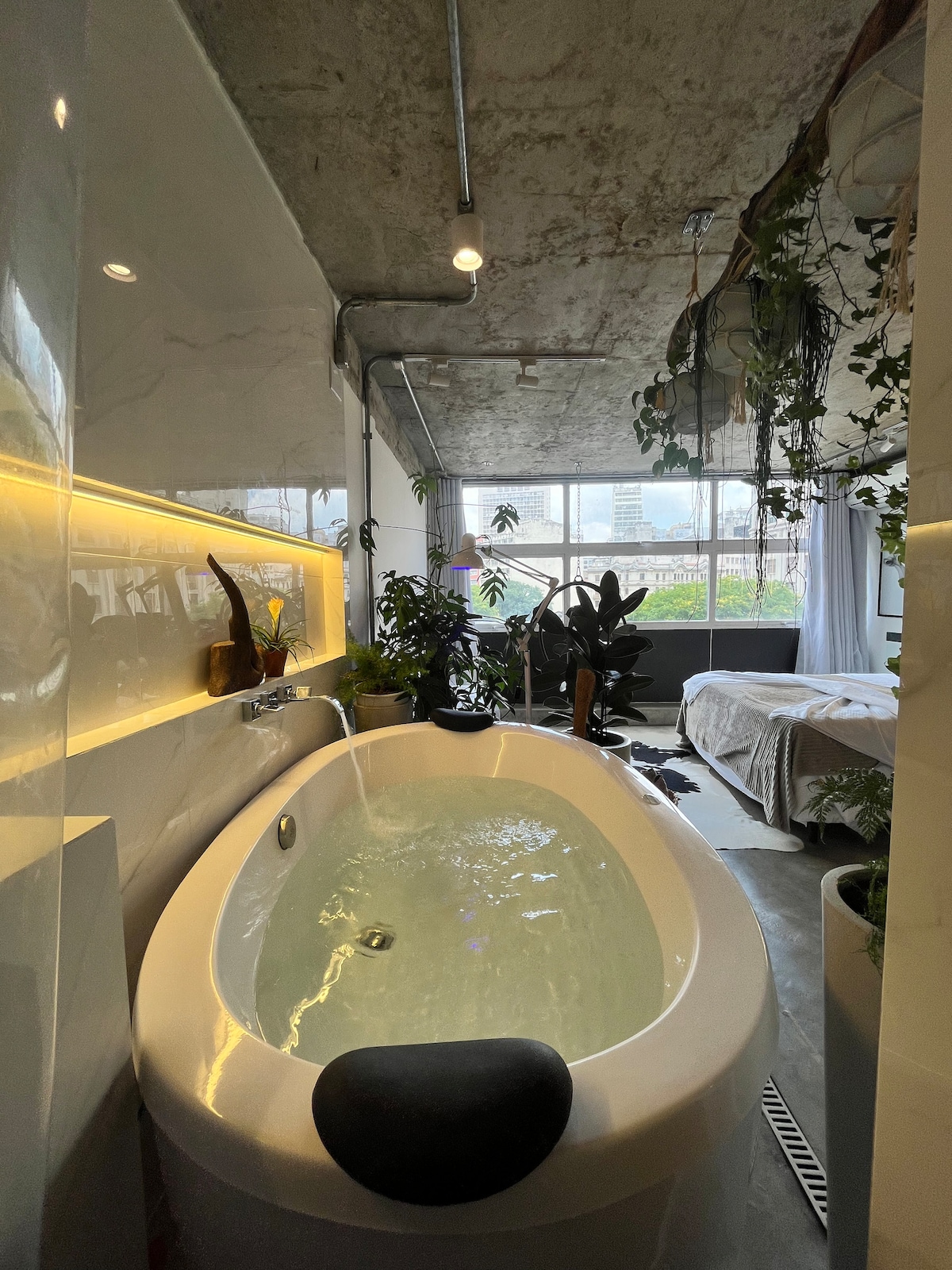 A spacious bathroom area is highlighted by a white freestanding tub filled with water, surrounded by lush green plants. Soft lighting enhances the relaxing ambiance, while large windows provide an urban view. The adjacent area features a neatly made bed and additional greenery.
