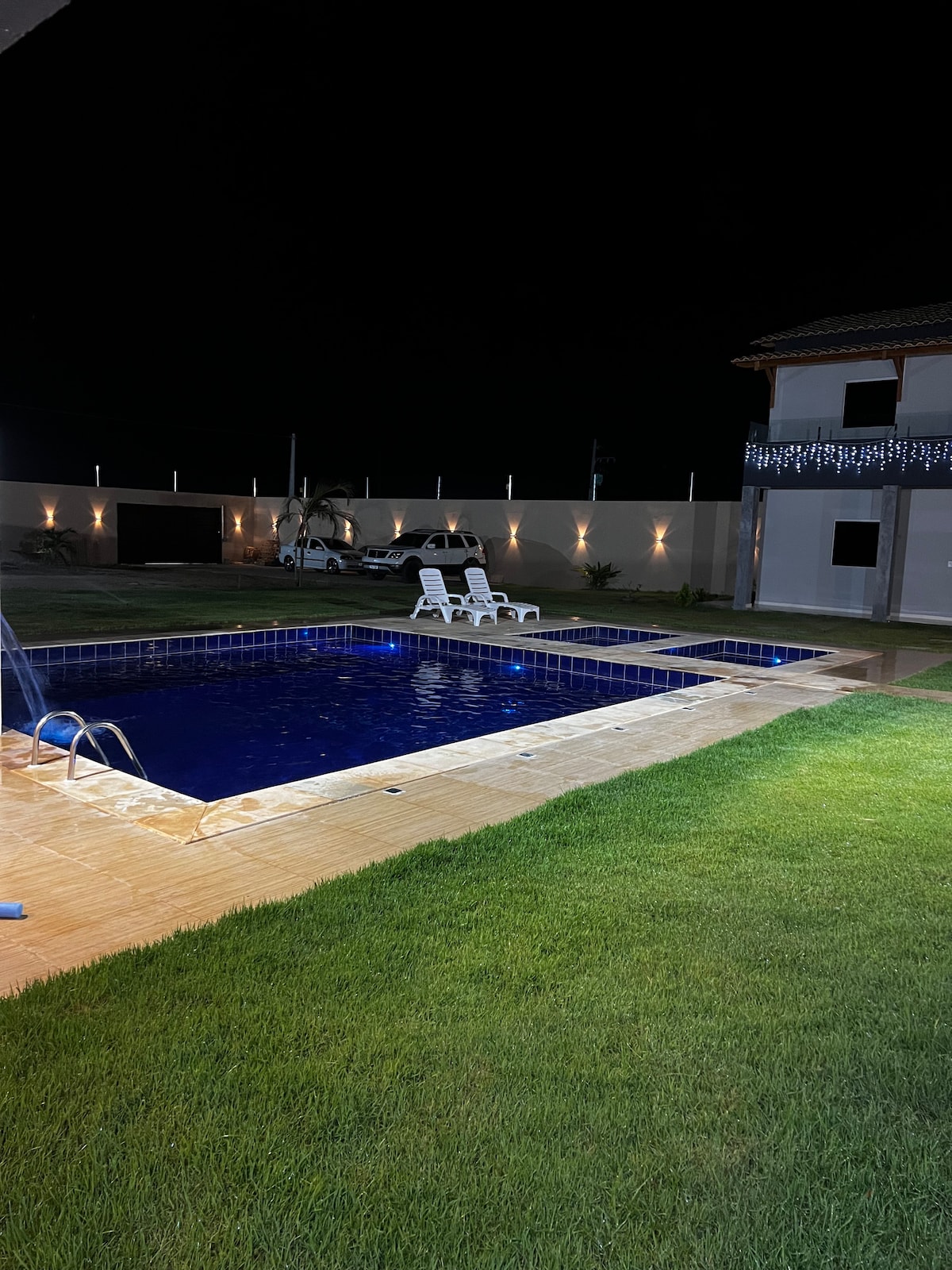 A well-lit outdoor area features a rectangular pool with a wooden deck and lounge chairs. The pool's water is illuminated, providing a serene atmosphere. A spacious lawn surrounds the area, with palm trees visible in the background, and the facade of a two-story building is on one side.
