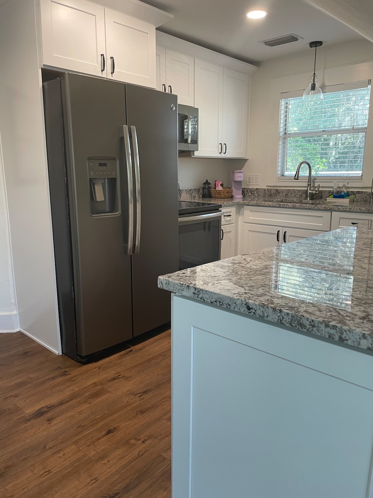 A modern kitchen is highlighted, featuring granite countertops and sleek cabinetry. Stainless steel appliances, including a refrigerator with a water dispenser, are visible. A window allows natural light to illuminate the space, while kitchen utensils and small appliances are organized neatly on the counters.