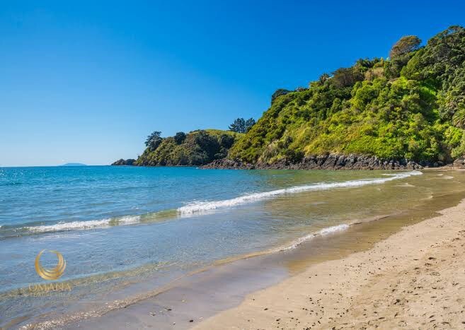 A tranquil beach scene showcases calm blue waters gently lapping against a golden sandy shore. Lush green hills rise in the background, framed by clear blue skies. The natural landscape invites relaxation and a connection with the coastal environment.