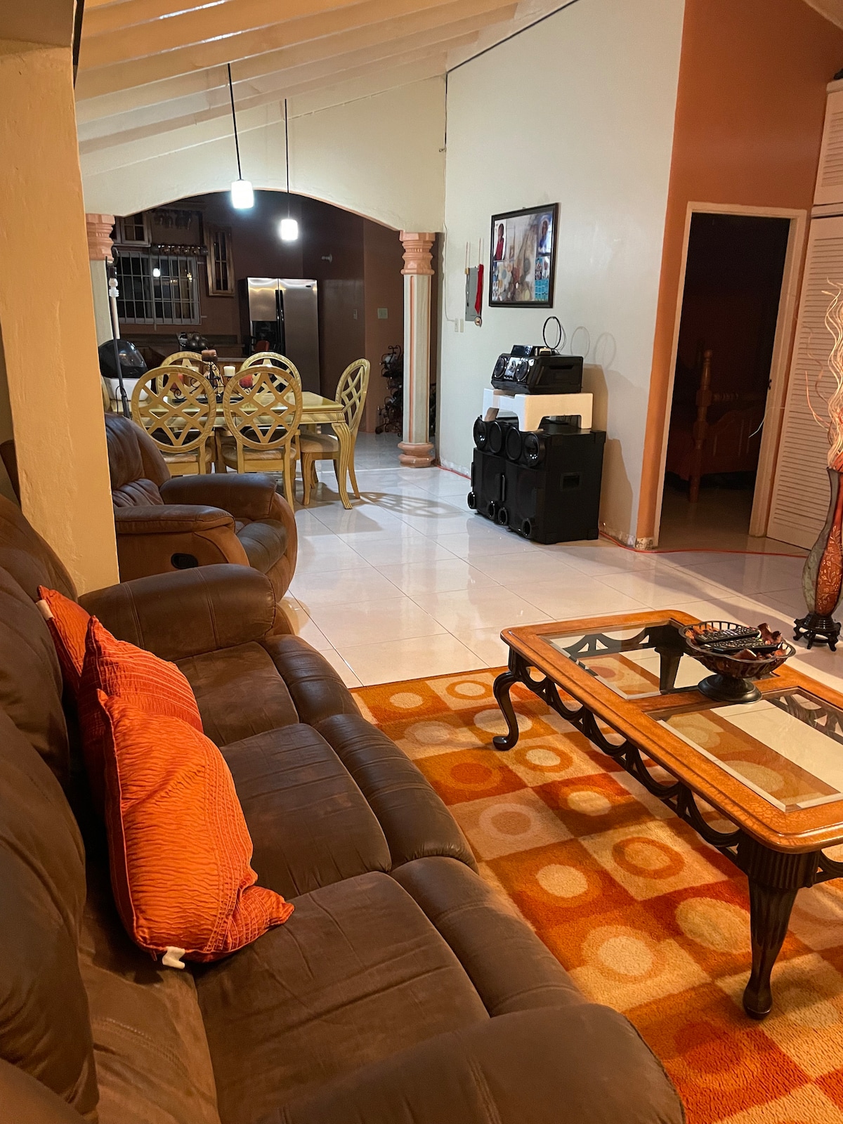 A comfortable living area features a dark brown couch adorned with orange cushions. A patterned area rug adds warmth under a glass coffee table. In the background, a dining space with a circular table and matching chairs is visible, along with an open kitchen design.