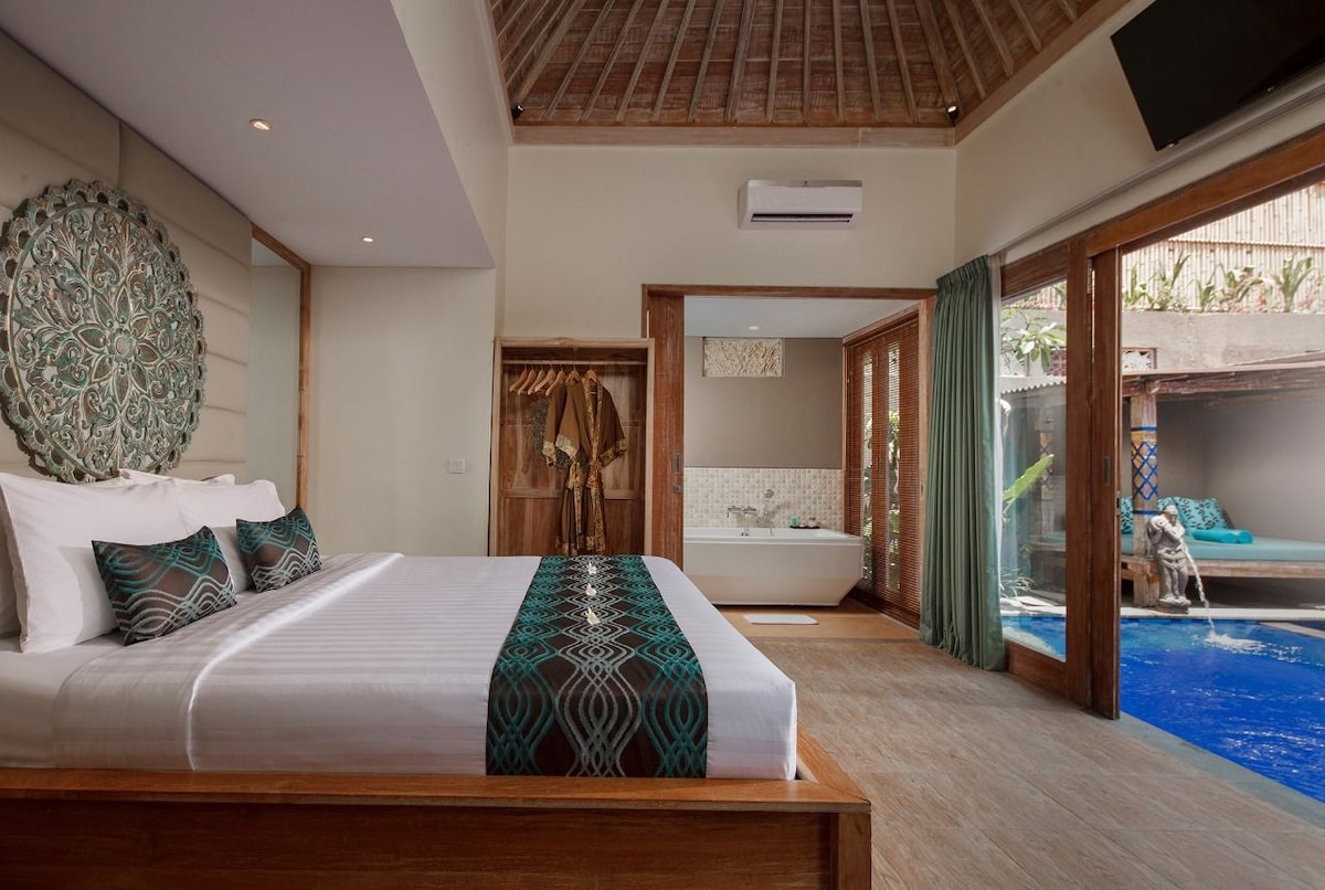 A spacious bedroom is featured, showcasing a large bed with a decorative throw across the top. Natural light filters through expansive glass doors, leading to a private outdoor area with a pool. A stylish wall hanging is visible, complementing the room's serene decor.