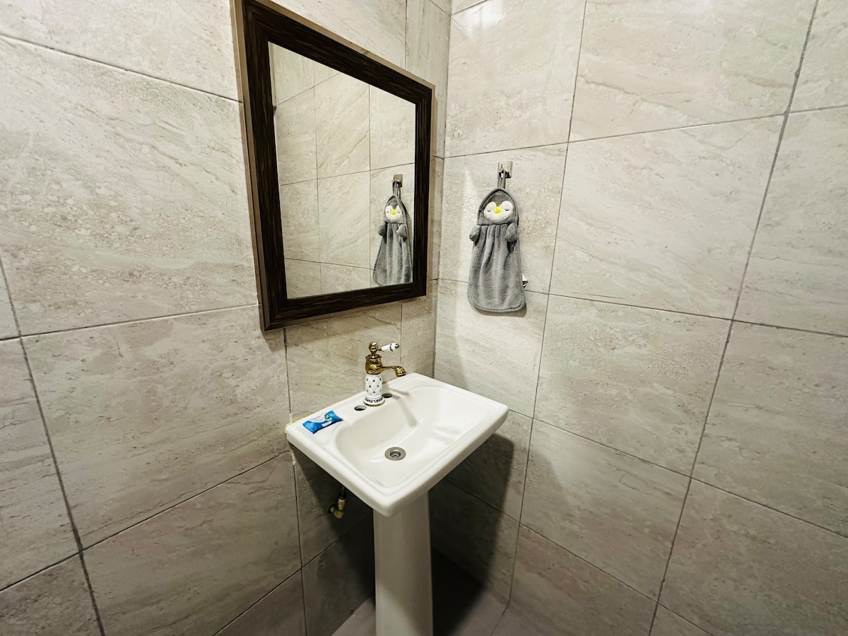 A clean and compact bathroom is depicted, featuring a white sink with a gold faucet. A large mirror framed in wood hangs above the sink. A towel with a character design is displayed on a hook, contrasting against the light stone tiles.