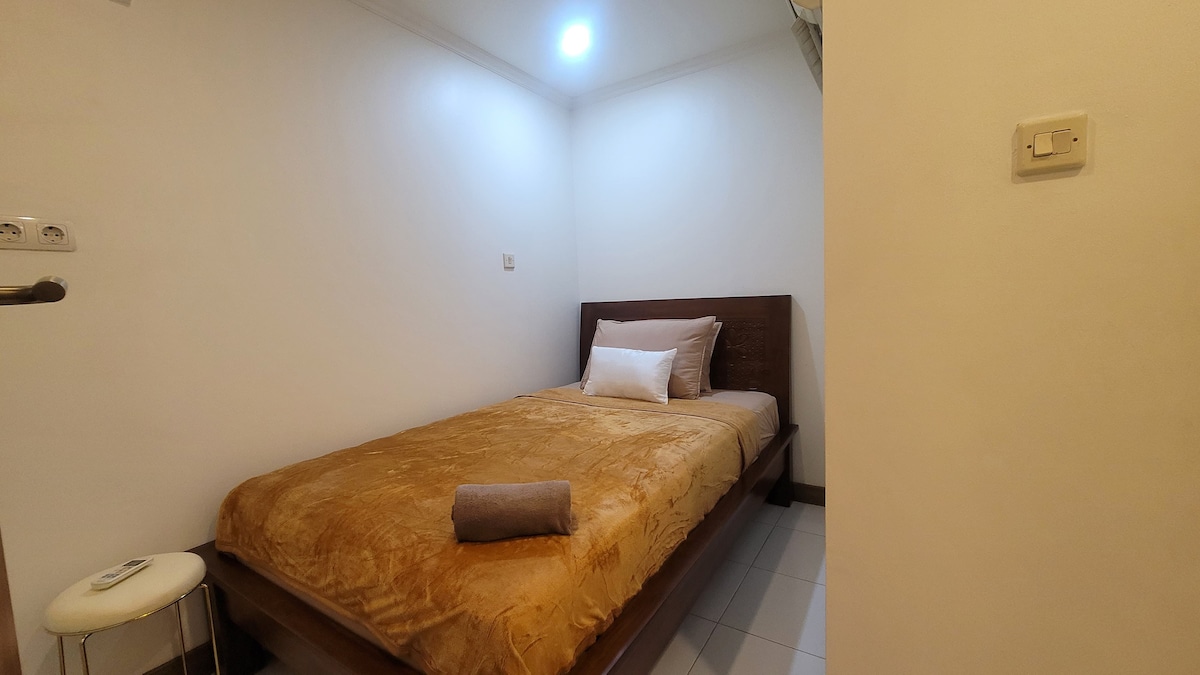 A sleeping area features a wooden bed with a soft beige blanket and a white accent pillow. A rolled towel is placed on the bed. A small white stool is positioned beside the bed. Soft lighting fills the room, which is finished in neutral tones.