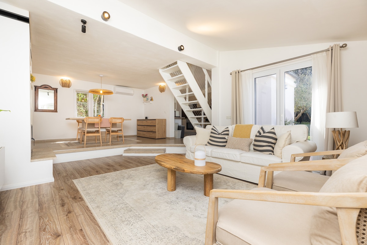 A light-filled living area features a comfortable white sofa adorned with decorative pillows. Light wood accents complement the space, which includes a wooden coffee table and a soft area rug. A dining area with a wooden table and chairs is visible in the background.