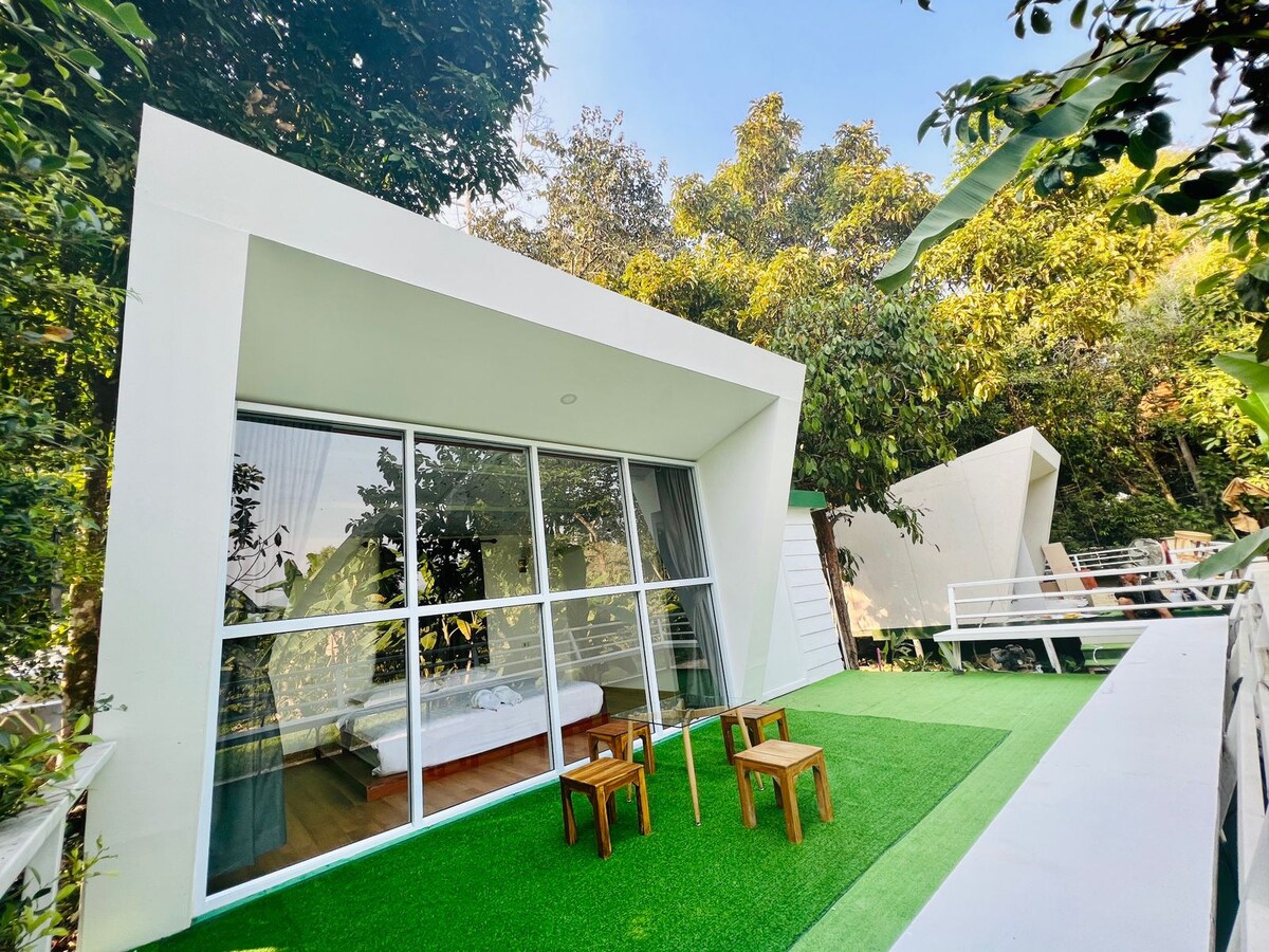 A modern structure features large glass windows, providing ample natural light to the interior. Outside, a small patio area is furnished with two wooden chairs and a side table, surrounded by lush greenery. The space is complemented by a well-maintained artificial lawn.