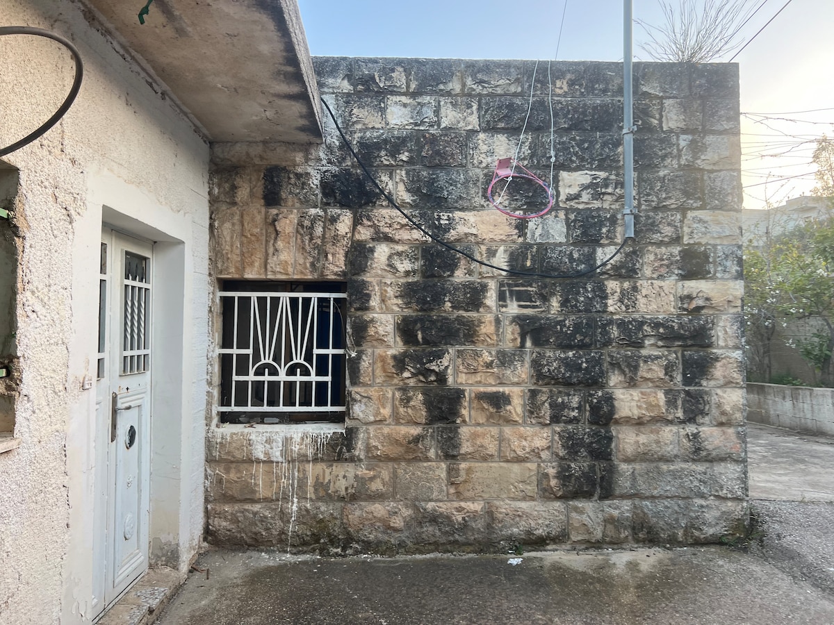 A stone wall is presented, featuring a window with a decorative metal grille. Nearby, a white door is visible, leading into the structure. Electrical wires hang overhead, and a certain rustic charm is conveyed by the weathered surfaces.