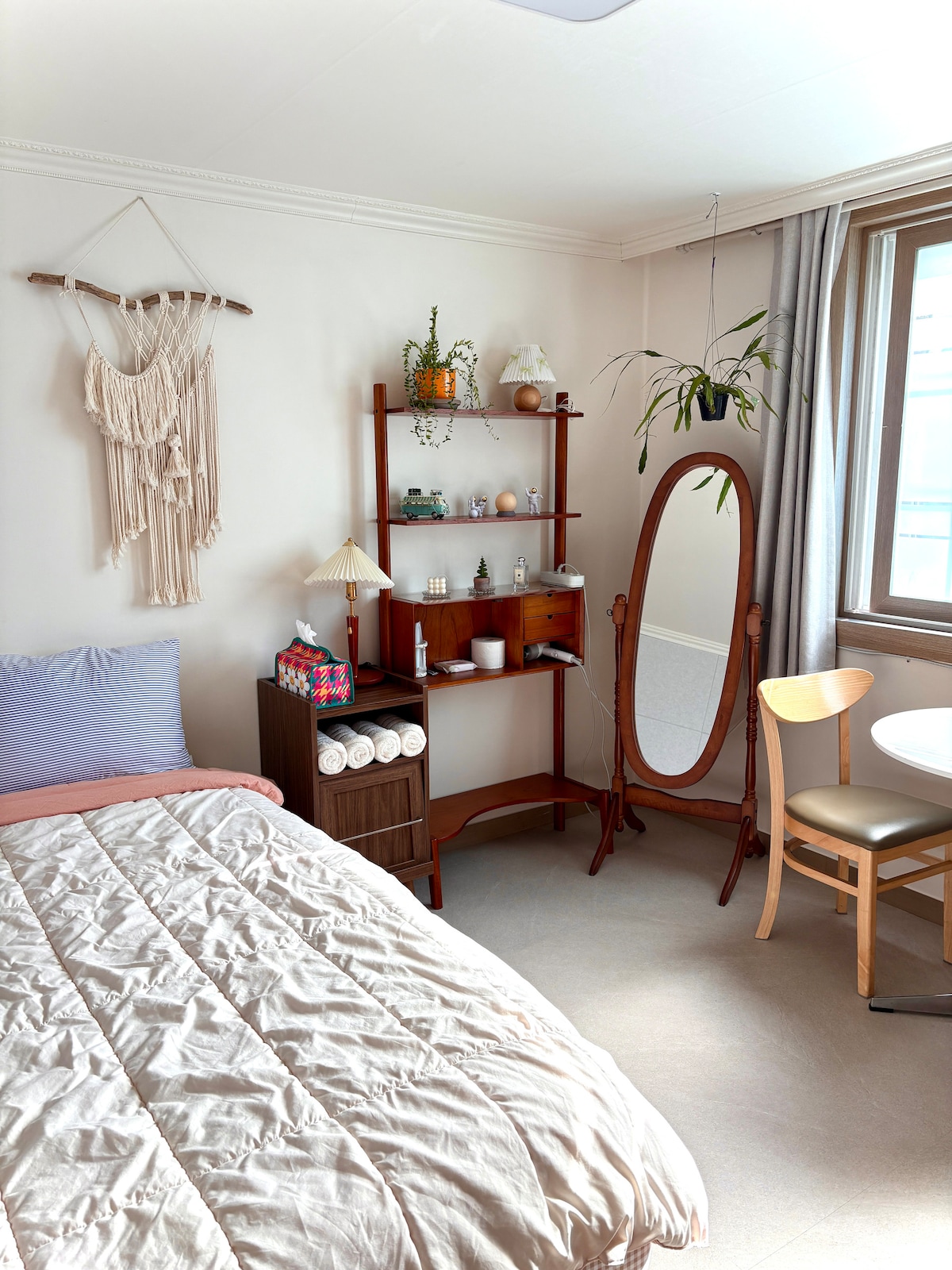 A cozy room is furnished with a spacious bed draped in a light quilt and a wooden shelf displaying decorative items. A stylish mirror stands nearby, accompanied by a chair and small table. Natural light filters through the curtains, illuminating the neutral-toned decor.