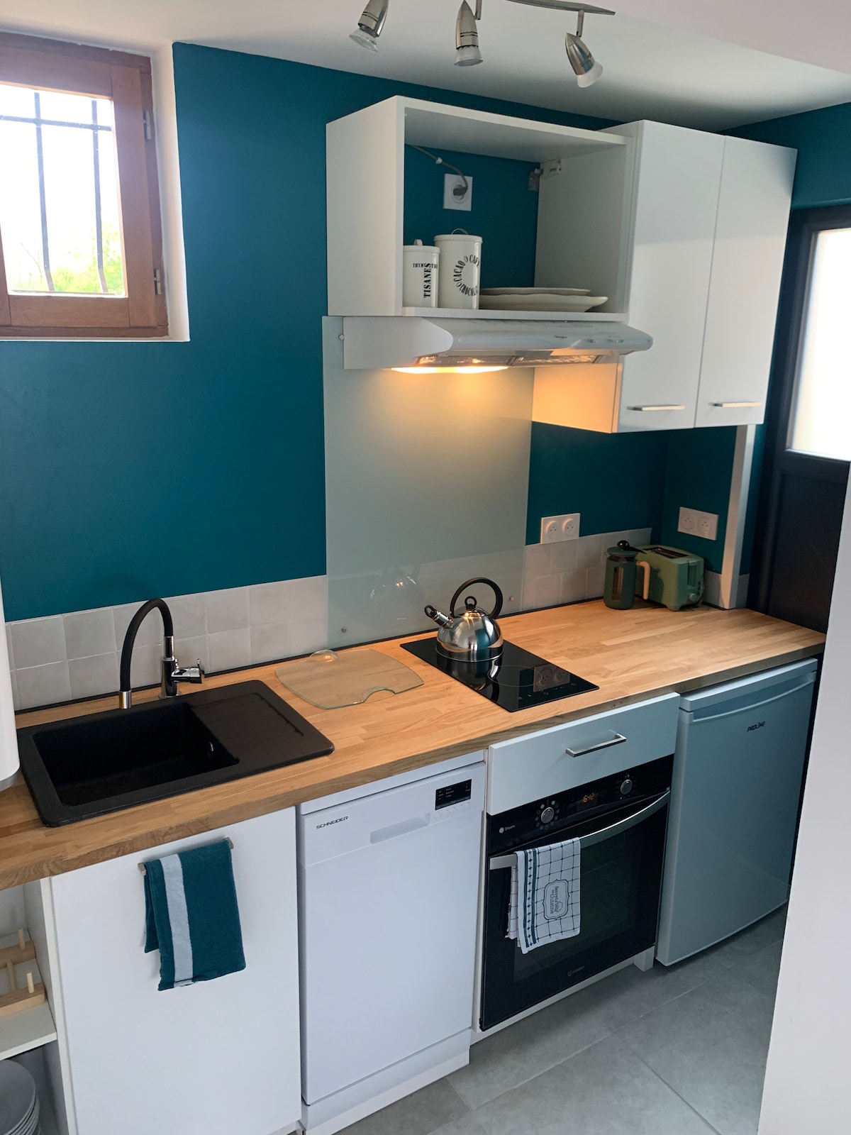 A compact kitchen features modern appliances including a stove, dishwasher, and refrigerator. A wooden countertop provides workspace, complemented by a sleek black sink. A kettle sits on the stovetop, and light streams through a nearby window, illuminating the teal accent wall.