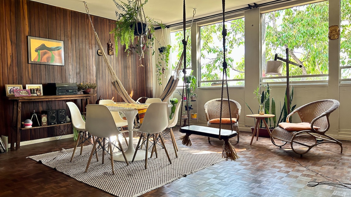 A spacious living area features a mix of seating options, including hanging swings and woven chairs. A large round table is surrounded by modern white chairs. Artwork is displayed on the wooden wall, while greenery from the window adds a refreshing touch to the space.