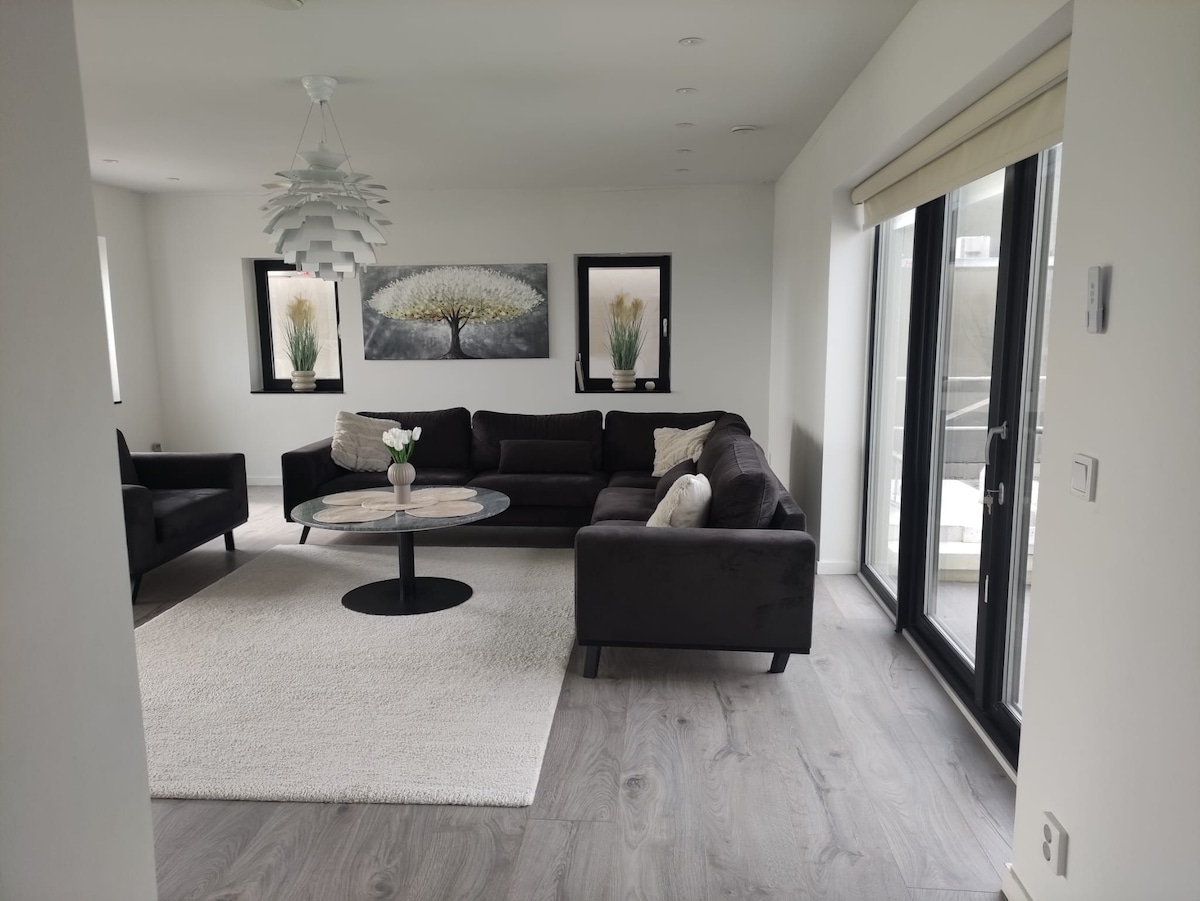 A modern living room features a dark sectional sofa and two black armchairs surrounding a circular coffee table. Tall windows provide natural light, with light-colored drapes partially drawn. Artwork with floral themes adorns the walls, and a plush area rug adds comfort underfoot.