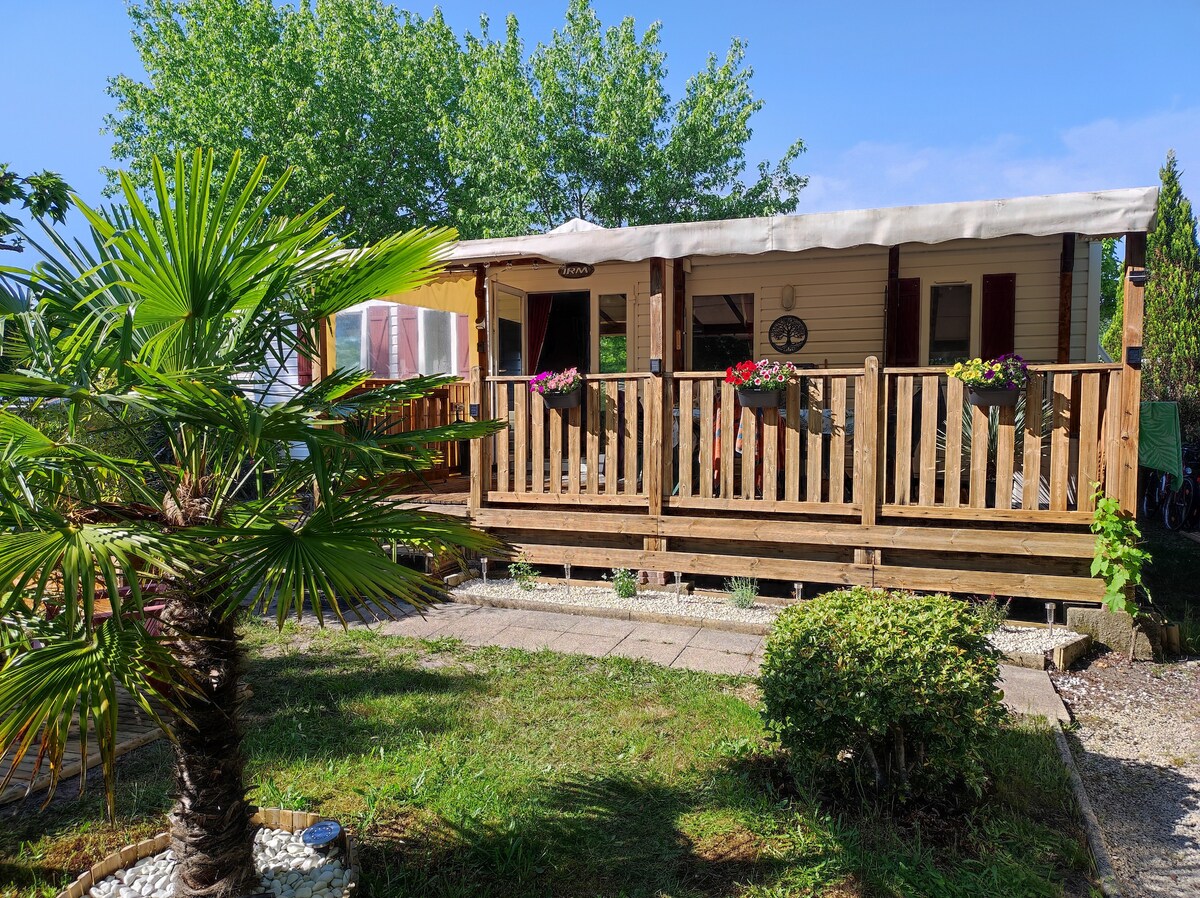 A mobile home is featured with a wooden deck surrounded by lush greenery. Potted plants with vibrant flowers adorn the exterior. The covered terrace provides shaded seating space and adds to the welcoming ambiance of the outdoor area.