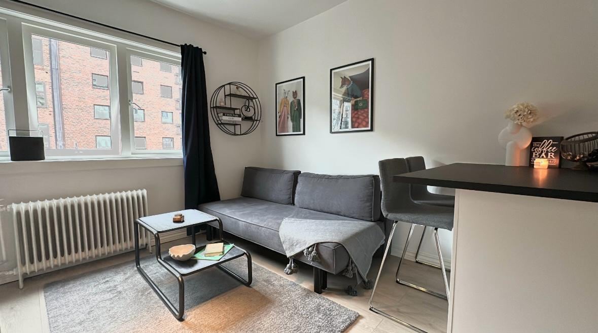A compact living area is highlighted by a gray sofa and a low coffee table, positioned on a textured rug. Two framed artworks are displayed on the light-colored walls, while natural light enters through large windows with dark curtains. A bar seating area is visible in the background.