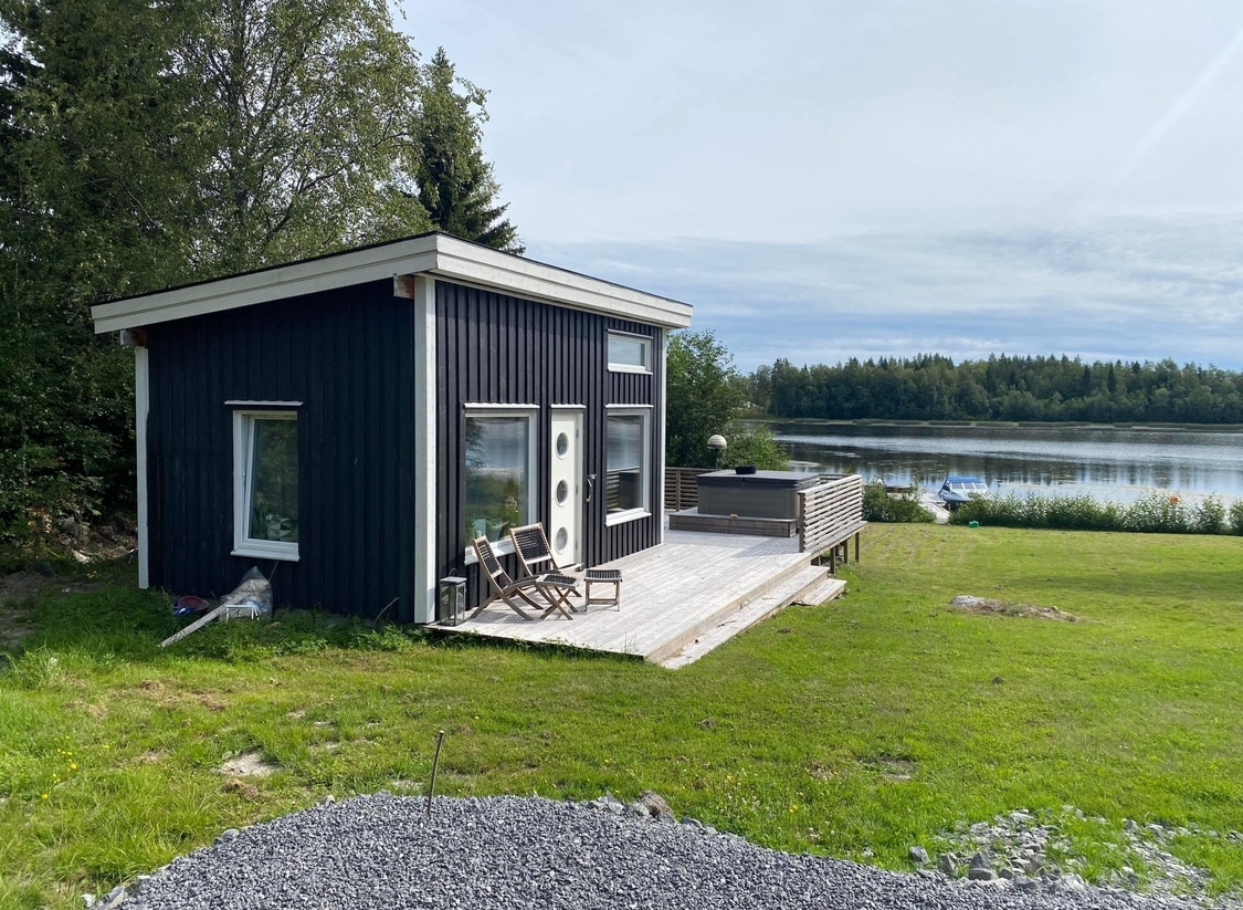 A contemporary black wooden house is situated near the water, showcasing large windows that provide a view of the serene landscape. A wooden deck features lounge chairs, and a well-maintained green lawn offers space for outdoor activities.