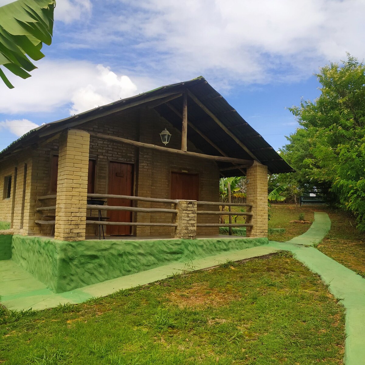 A charming building with a rustic design features a broad porch supported by wooden beams. The path leading up to the entrance is lined with greenery, enhancing the natural setting. The structure is surrounded by lush vegetation and a clear blue sky.