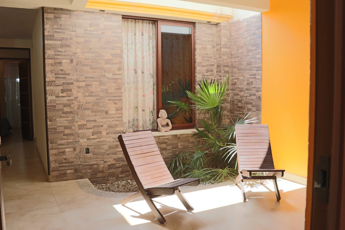 A serene outdoor area is featured, showcasing two wooden lounge chairs positioned on a light-colored tiled floor. Natural light filters through a nearby window, illuminating the space that includes a decorative plant and a stone border, creating a calming environment.