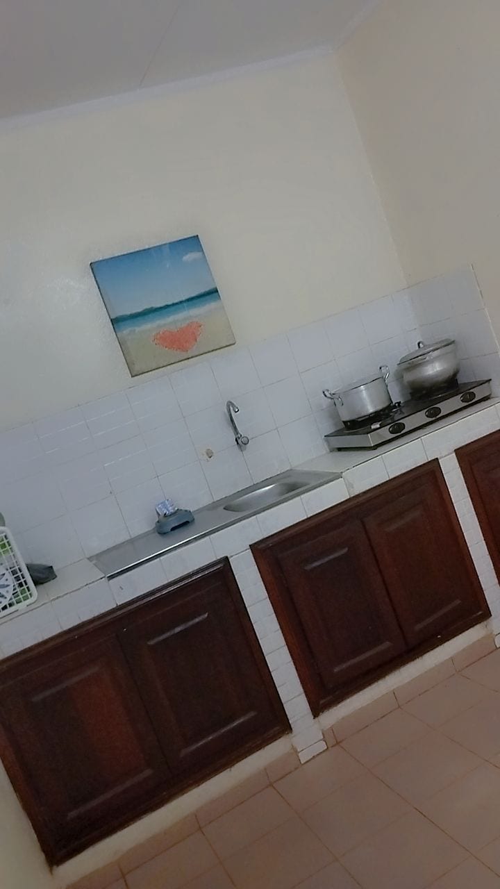 A practical kitchen space is equipped with wooden cabinetry and a white tiled backsplash. A small sink and a two-burner stove are visible, alongside cooking pots. A beach-themed painting adds a touch of color to the otherwise neutral walls.