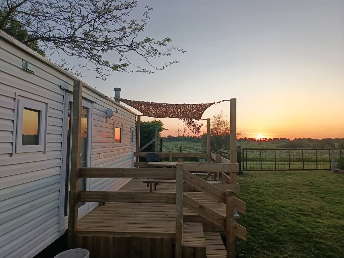 A mobile home with a wooden deck is pictured at sunset, featuring a shaded seating area beneath a woven covering. Green fields extend in the background, blending into the orange and purple hues of the evening sky.
