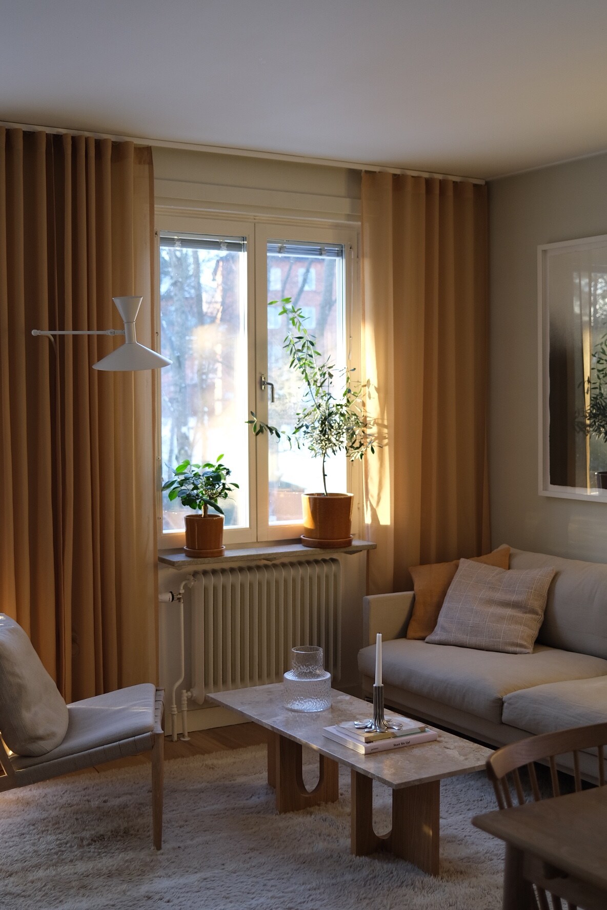 A bright living room is illuminated by natural light filtering through a window adorned with simple curtains. Two potted plants are positioned on the sill, enhancing the space. A light-colored sofa is complemented by a cozy chair, with a wooden coffee table at the center.