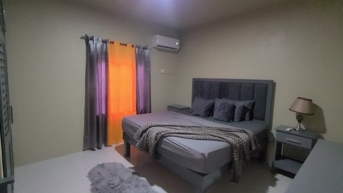 A spacious bedroom is featured with a large grey bed adorned with decorative pillows and a textured blanket. Soft curtains in shades of grey and orange frame a window. An air-conditioning unit is positioned above, and a bedside table holds a lamp.