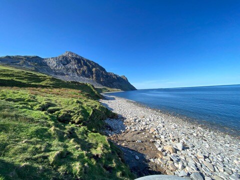 A rugged shoreline is framed by gentle green hills, leading to a dramatic rocky mountain rising sharply against a clear blue sky. The calm sea reflects the sky's hues, creating a serene coastal scene. Smooth pebbles form the beach, gradually merging with the water's edge.