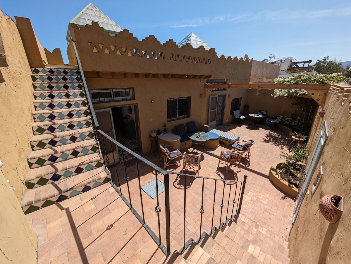 A courtyard is depicted, featuring earthy tones and terracotta tiles. Comfortable seating arrangements are visible, including sofas and chairs, with a round table at the center. Stairs lead to a higher level, framed by decorative railings. Lush greenery adds a natural element to the space.