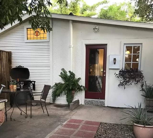 The exterior of the apartment features a welcoming entrance with a red door. A small patio area is shown with a bistro set, surrounded by potted plants. A barbecue grill is placed nearby, enhancing the outdoor living space.