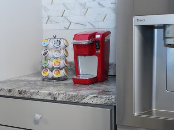 A red coffee maker is positioned on a speckled grey countertop. A rotating holder with various coffee pods sits beside it, showcasing a selection of flavors. The background features a marble backsplash that enhances the kitchen's modern aesthetic.