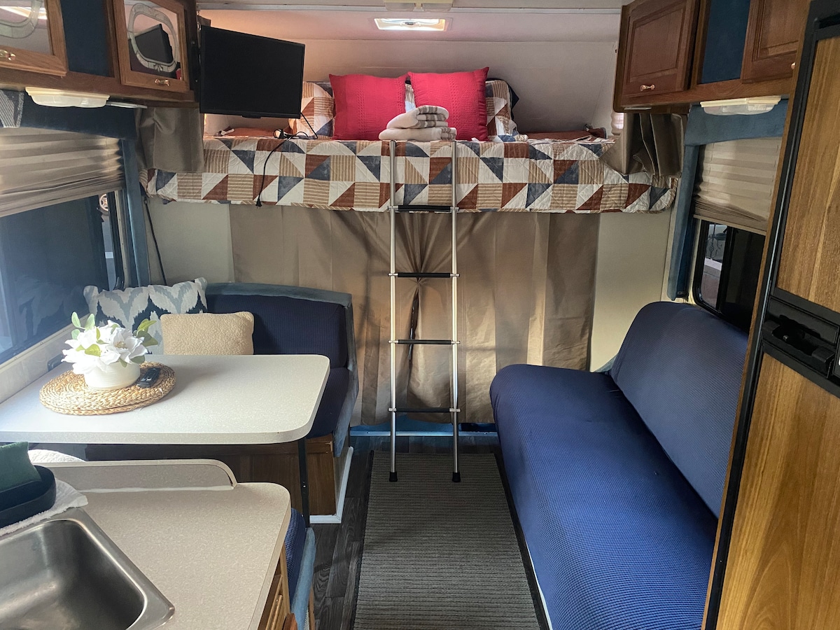 The interior of the RV features a seating area with a blue couch and a dining table. A ladder leads to a raised sleeping area above, which is adorned with a patterned quilt. A small television is mounted near the sleeping space.