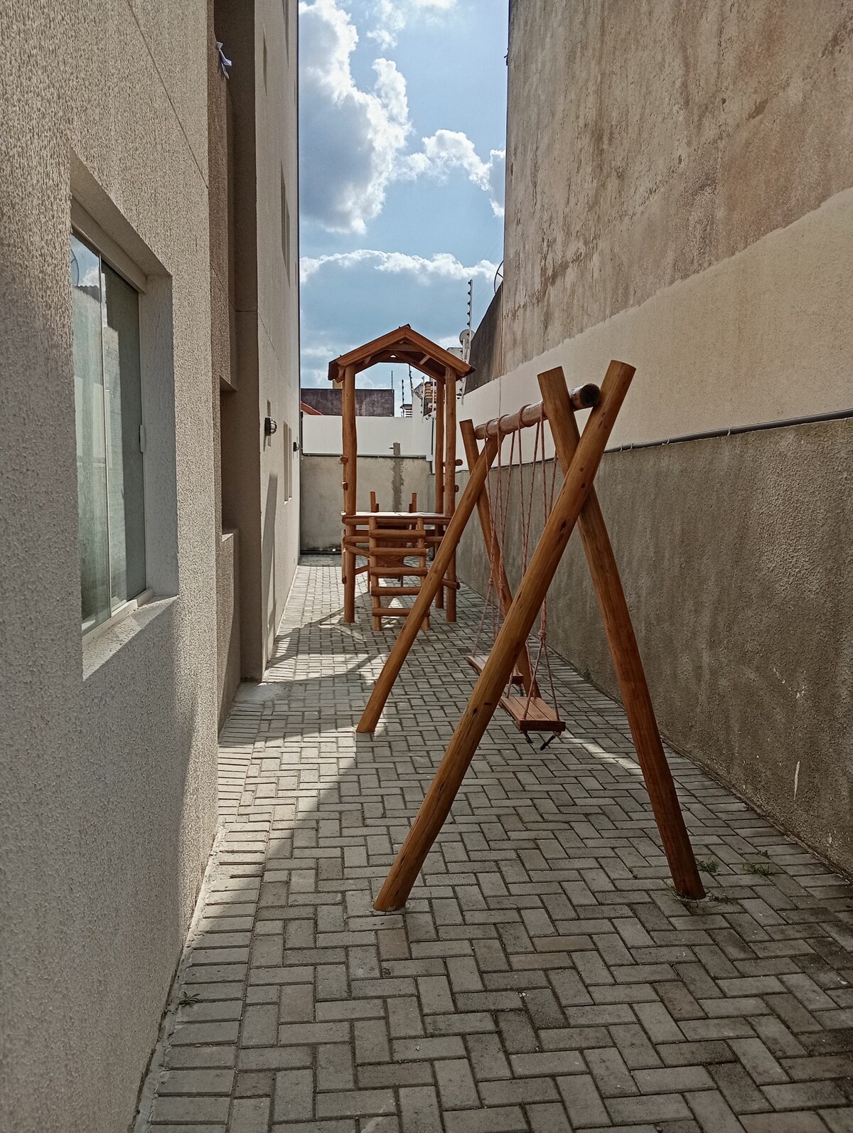 An outdoor play area is visible, featuring a wooden swing set and a climbing structure. The pathway, paved with bricks, runs alongside a building wall, and the sky is partially cloudy, providing gentle sunlight to the space.