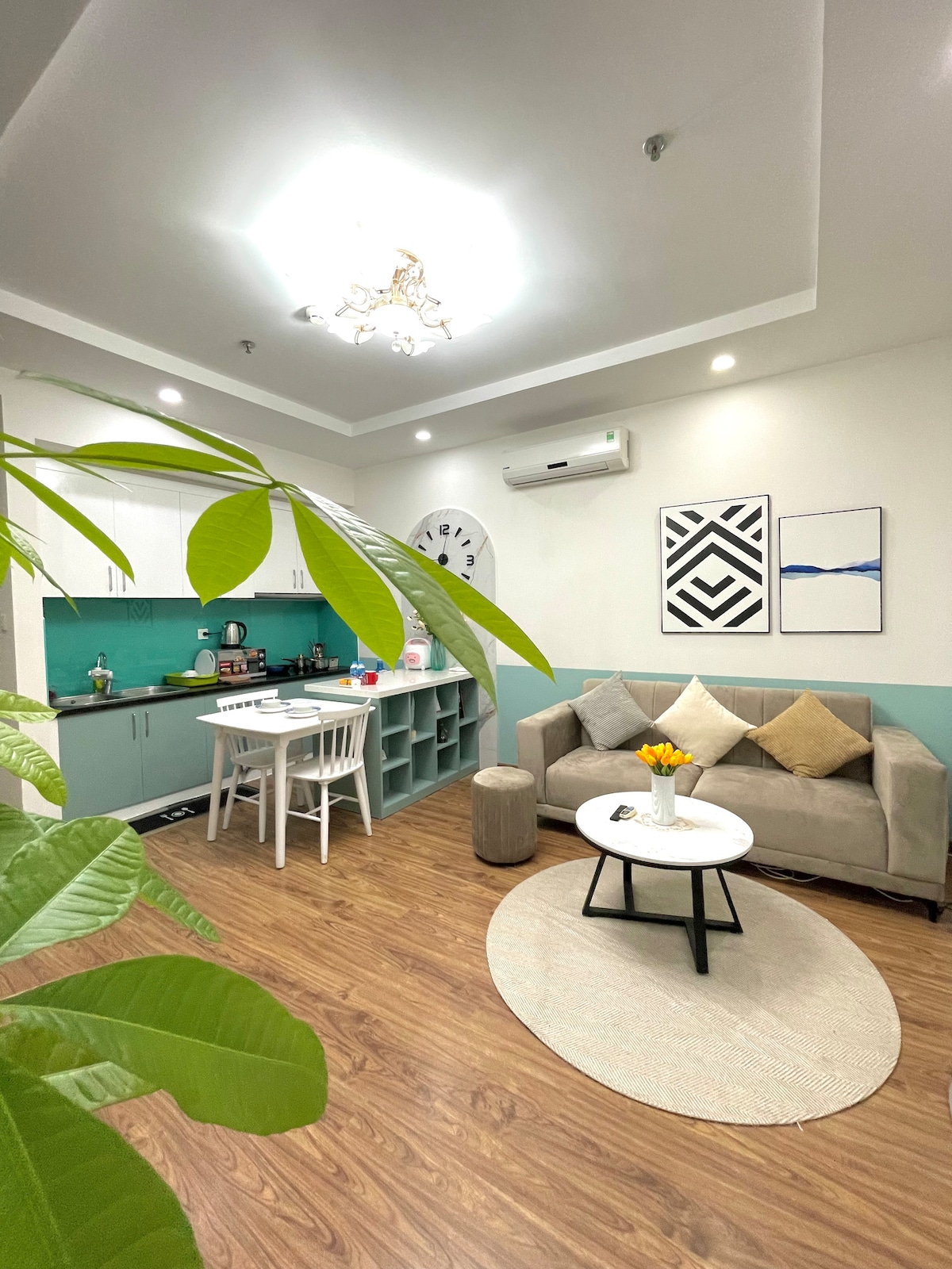 A well-appointed living area includes a comfortable sofa facing a round coffee table on a circular rug. A dining space with a white table and chairs is visible next to a teal kitchen area, enhanced by decorative plants and modern art pieces on the walls.