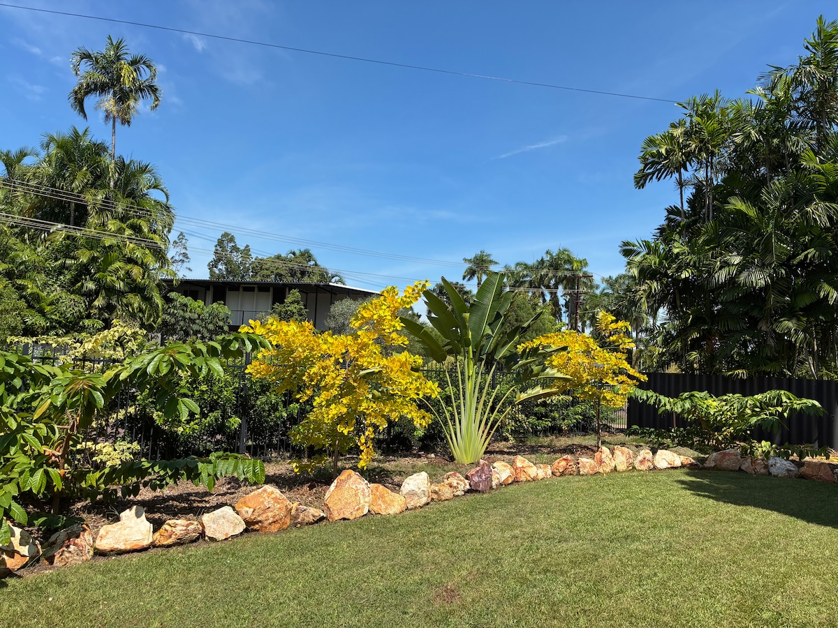 A lush garden area is featured, showcasing vibrant yellow flowering plants and tall green foliage. A rock border defines the garden, while a clear blue sky is visible above. The scene provides a sense of tranquility in the outdoor space.
