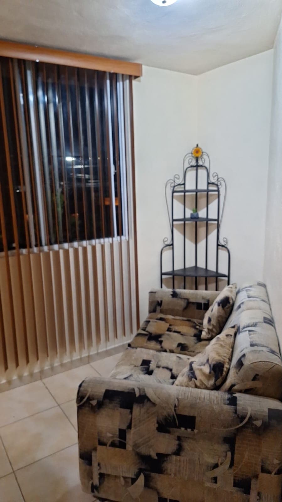 A cozy seating area features a patterned sofa positioned against a window dressed with vertical blinds. A metal shelf stands in the corner, showcasing decorative items, while natural light filters through the blinds, creating a warm ambiance.