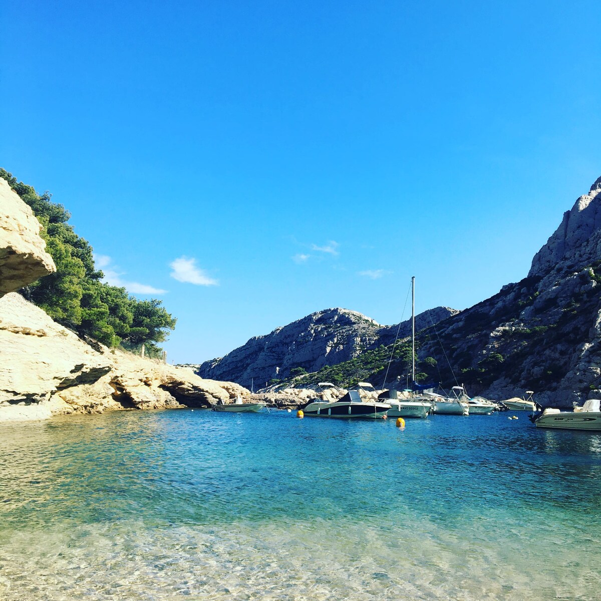 A serene cove features clear blue water surrounded by rocky cliffs and greenery. Several boats are anchored in the calm waters, and the bright sky enhances the tranquil setting. The shoreline is visible with smooth, sandy areas leading into the gentle waves.