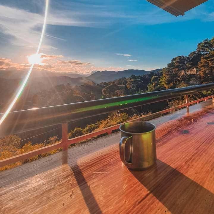 A scenic view of the sunrise over distant mountains is displayed. A stainless steel mug is positioned on a wooden railing, highlighting the tranquil outdoor space. Soft light illuminates the area, enhancing the natural beauty of the surrounding landscape.