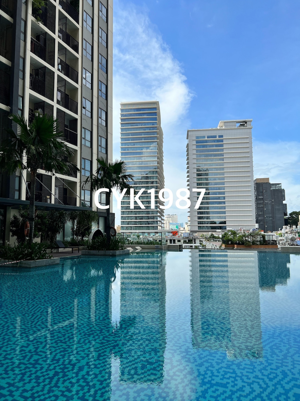 A tranquil swimming pool reflects the surrounding tall buildings against a blue sky. Palm trees line the pool's edge, contributing to a peaceful outdoor atmosphere within the apartment complex.