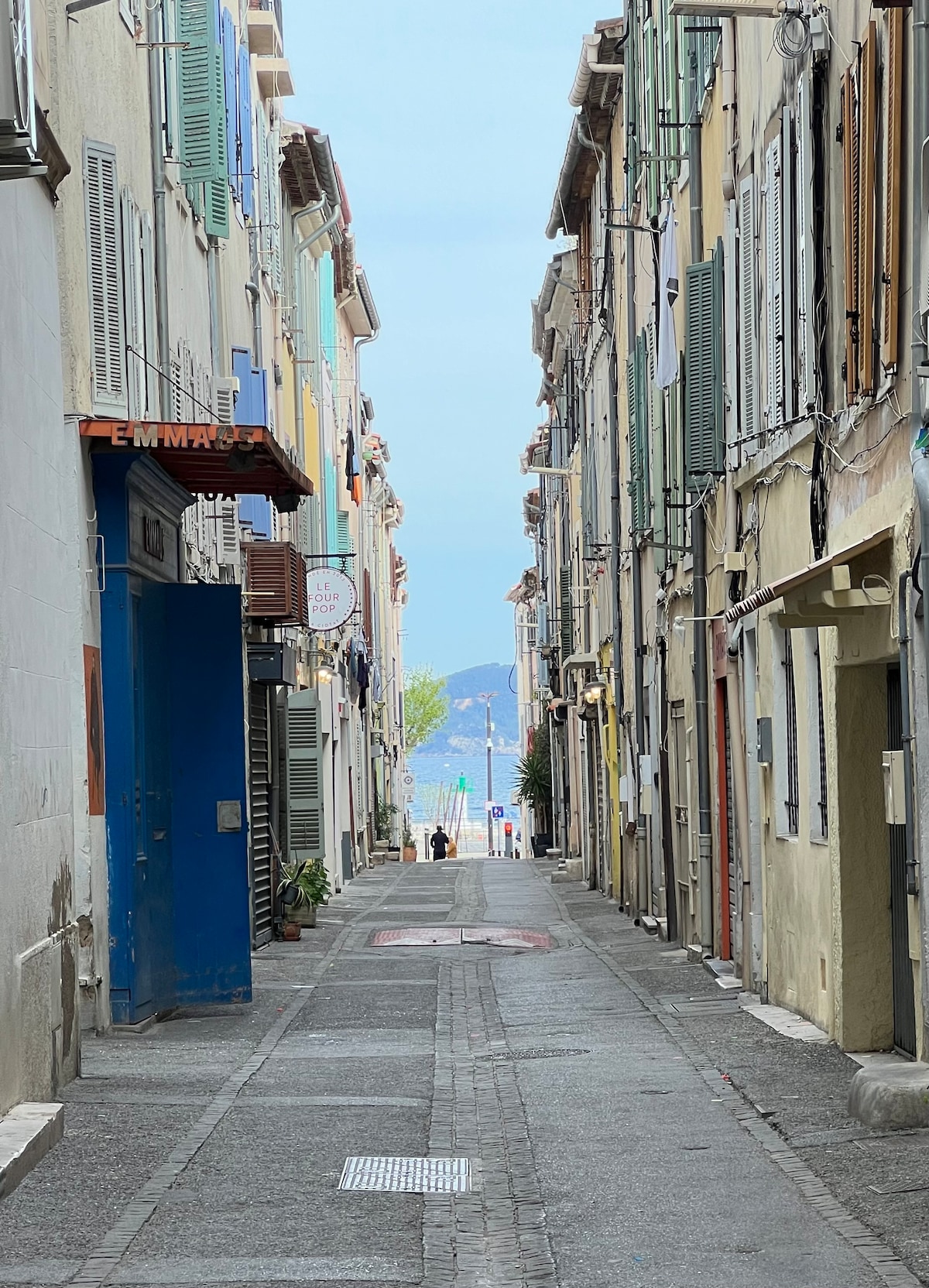 A narrow street lined with colorful buildings is shown, leading towards a view of the sea in the distance. The pathway is paved with stones, and a few people can be seen walking, adding life to the serene atmosphere of the charming location.