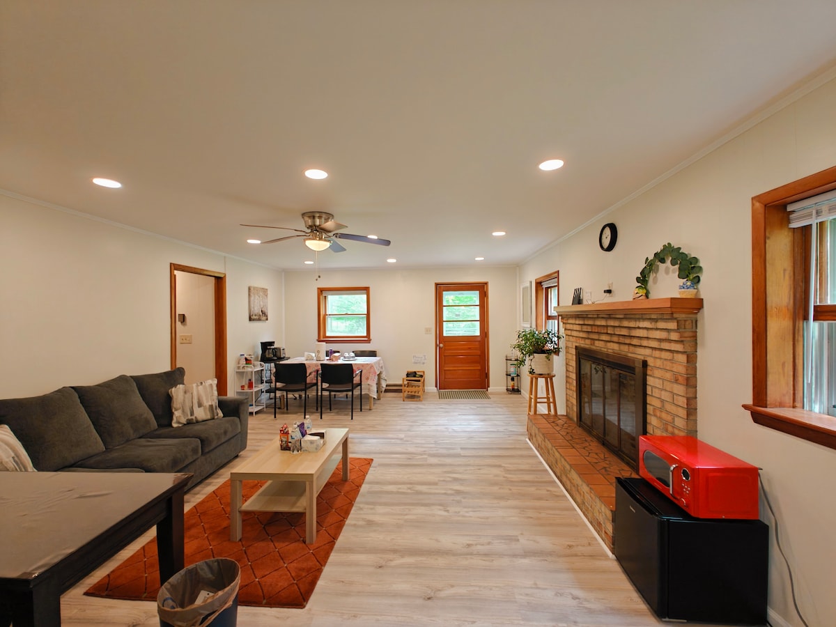 A bright living room features a cozy seating area with a dark sectional sofa. A coffee table sits on an area rug, while a brick fireplace serves as a focal point. Natural light streams through the windows, enhancing the spacious feel.
