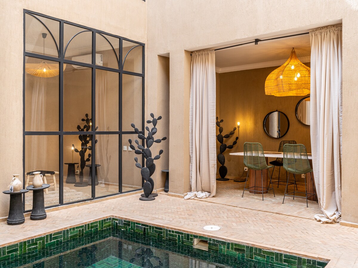 An inviting courtyard features a green-tiled pool surrounded by rustic tiles. A dining area with a light fixture hangs above a table, complemented by woven chairs. Decorative plants are placed intentionally along the courtyard walls, enhancing the serene setting.