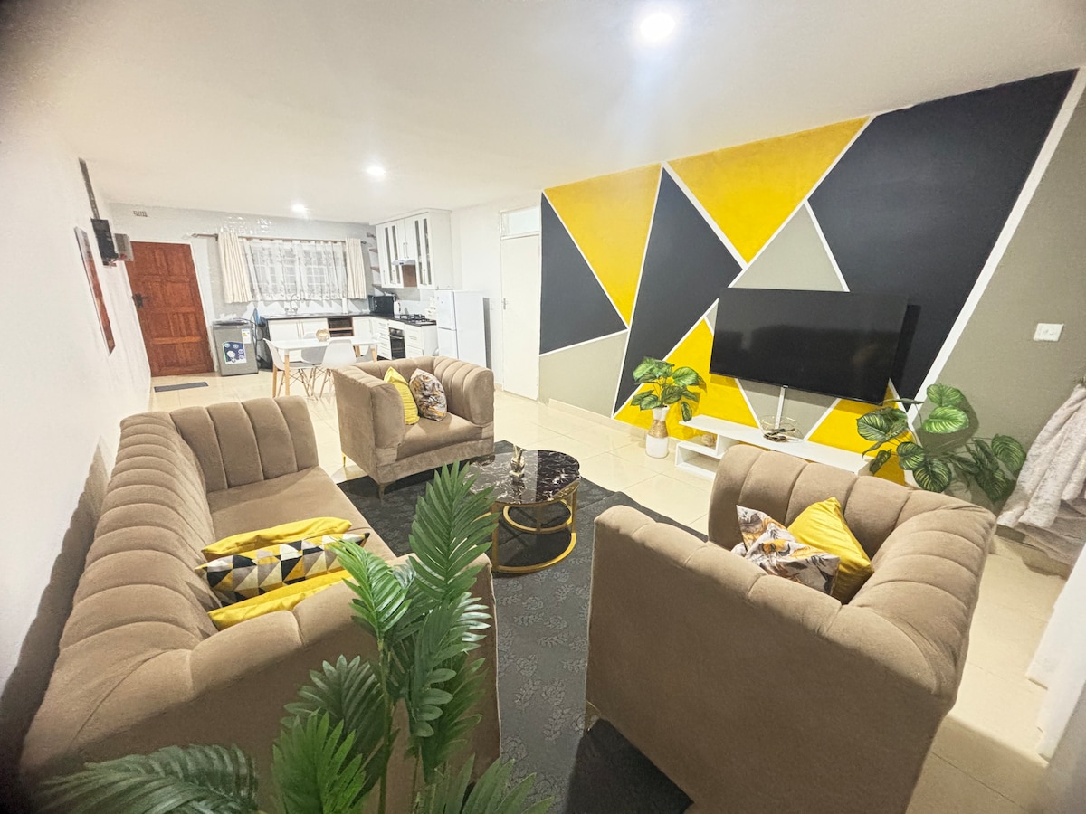 A modern living area features two beige sofas, accented with colorful cushions, arranged around a sleek coffee table. A bold geometric wall in yellow and black serves as a focal point, while lush green plants add a touch of nature to the space.