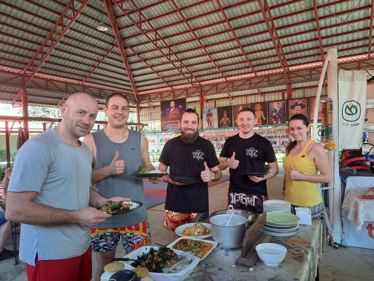 A group of individuals is standing together in a communal dining area. Plates filled with various dishes are held in their hands. A range of colorful food items are visible on the table, with a spacious and open arena in the background, accented by vibrant artwork on the walls.