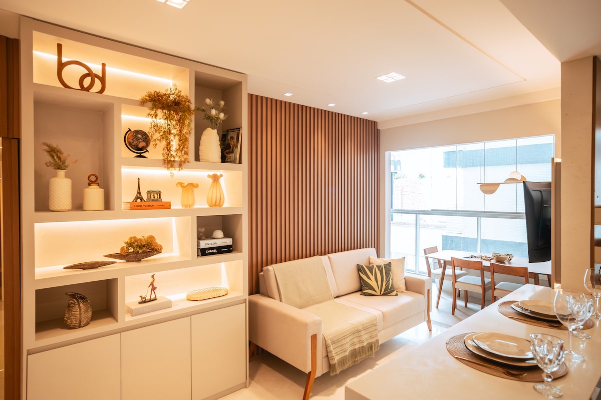 A contemporary living area is highlighted by a stylish shelving unit featuring various decorative items. A comfortable sofa is positioned near large windows, allowing natural light to fill the space. A dining table with chairs can be seen in the background, enhancing the open layout.