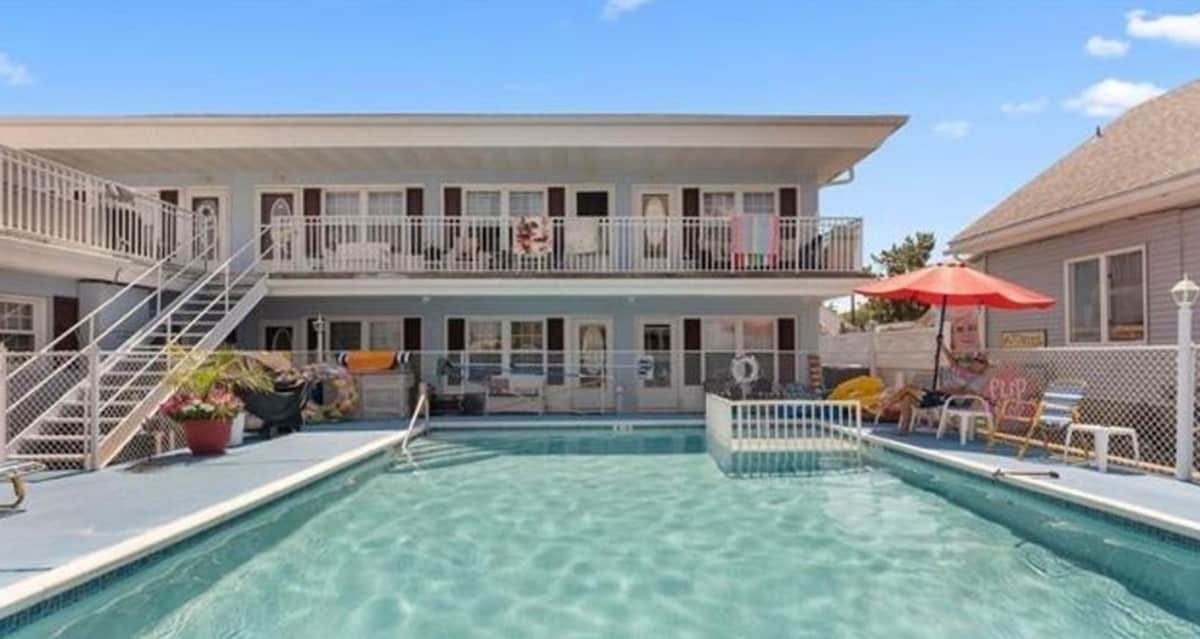 A shared heated pool is surrounded by a sunbathing area with lounge chairs and an umbrella, offering a relaxing environment. The two-story building features multiple entrances and a balcony with chairs, providing access to the outdoor spaces for guests.