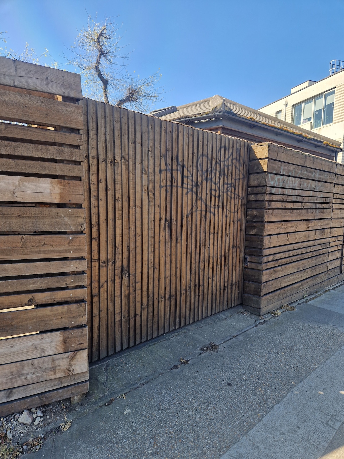 A wooden gate, made of slatted planks, is set within a surrounding wooden fence that offers privacy. The sky above is clear and blue, indicating a bright day. The gate is unassuming, with subtle graffiti markings on the wood.