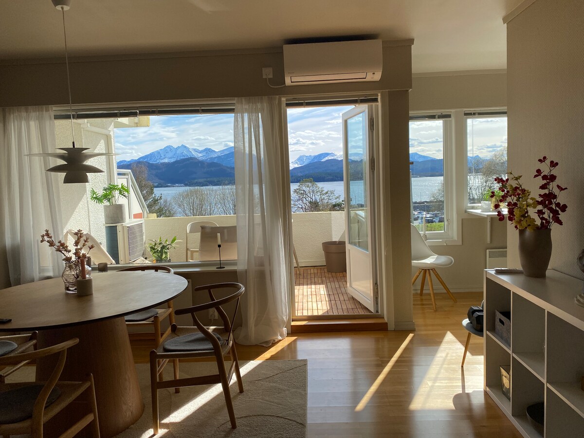 The living area presents large windows that offer expansive views of the fjord and surrounding mountains. A wooden dining table is positioned in the foreground, complemented by chairs and a decorative vase atop it. Natural light fills the space, creating a bright atmosphere.