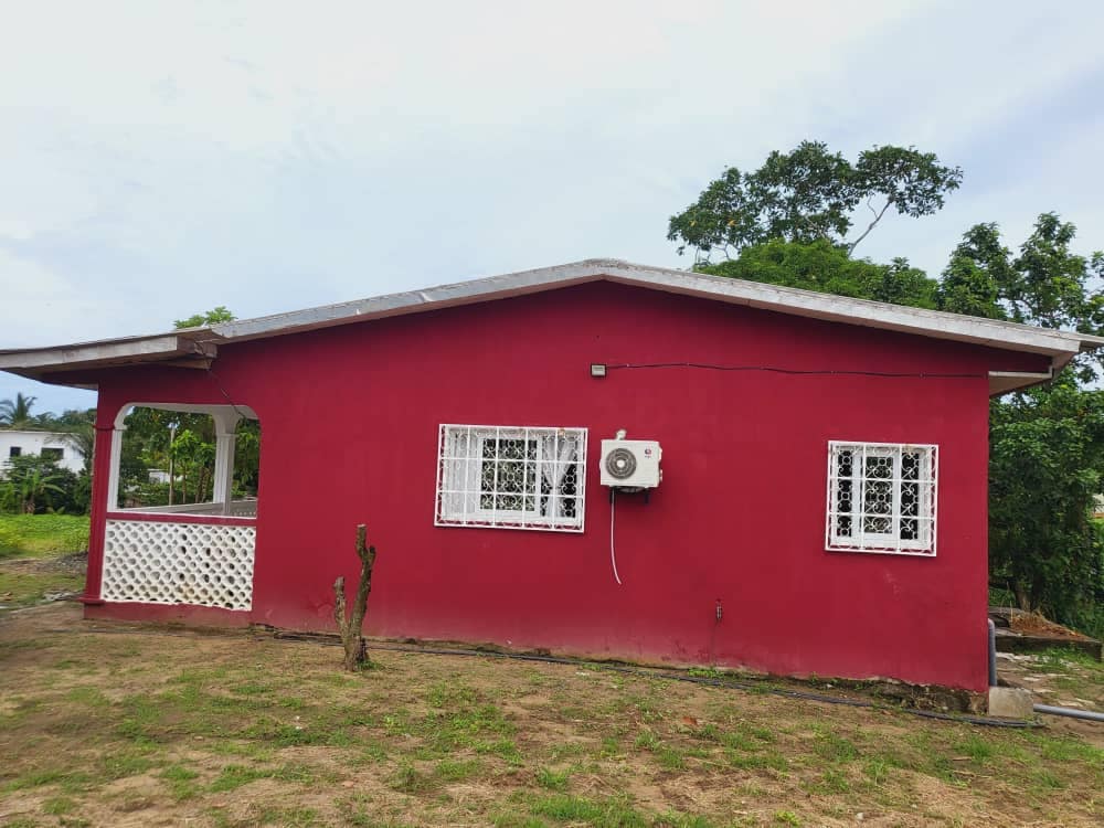 The exterior of the villa is shown, featuring vibrant red walls and white window frames that provide contrast. A small air conditioning unit is visible on the side. Surrounding greenery adds to the natural setting, enhancing the overall ambiance of the location.
