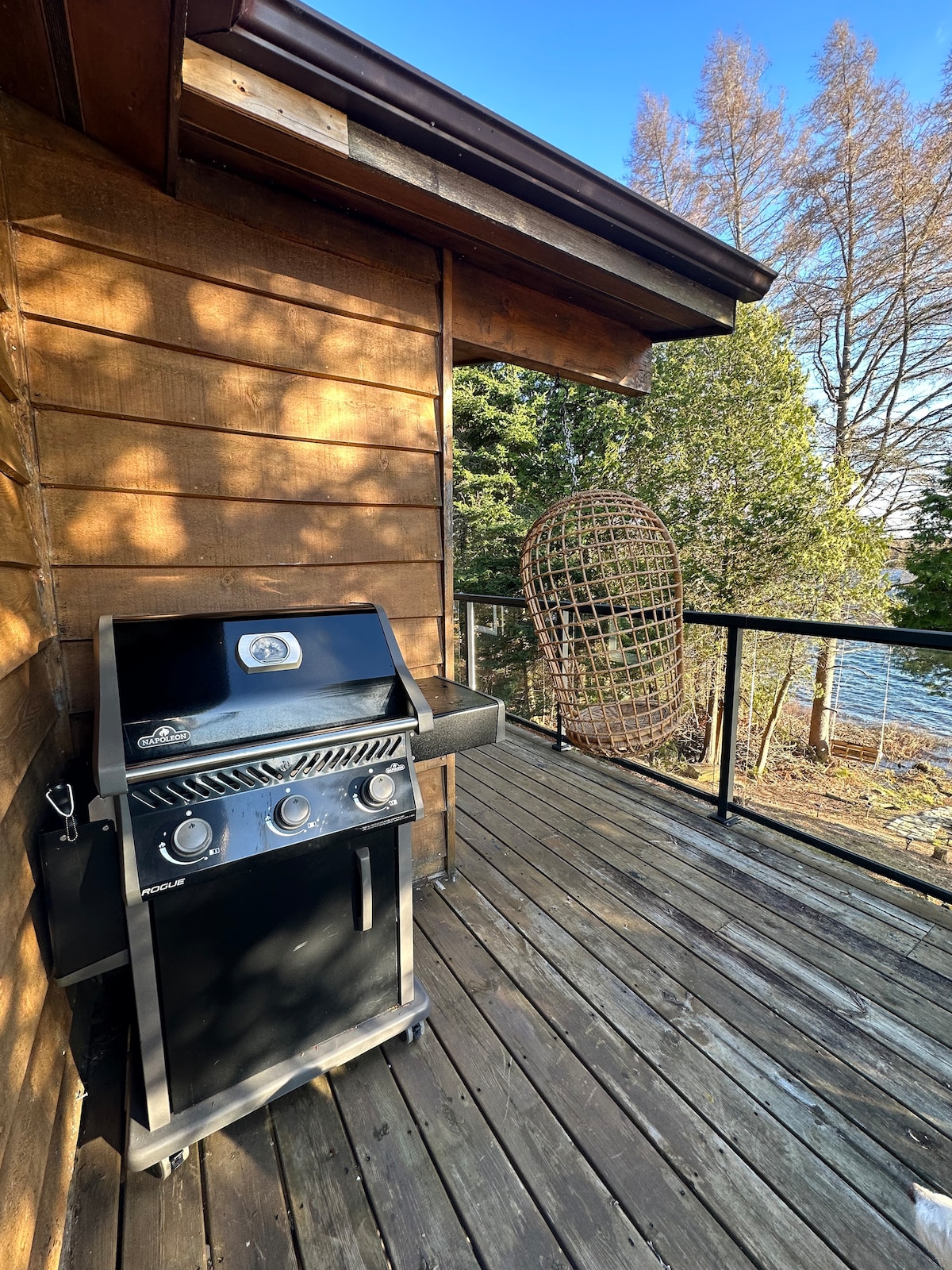 A black gas grill is positioned on a wooden deck, accompanied by a cozy hanging chair. The view features lush trees lining the edge of a serene lake, providing a tranquil outdoor space ideal for gatherings.
