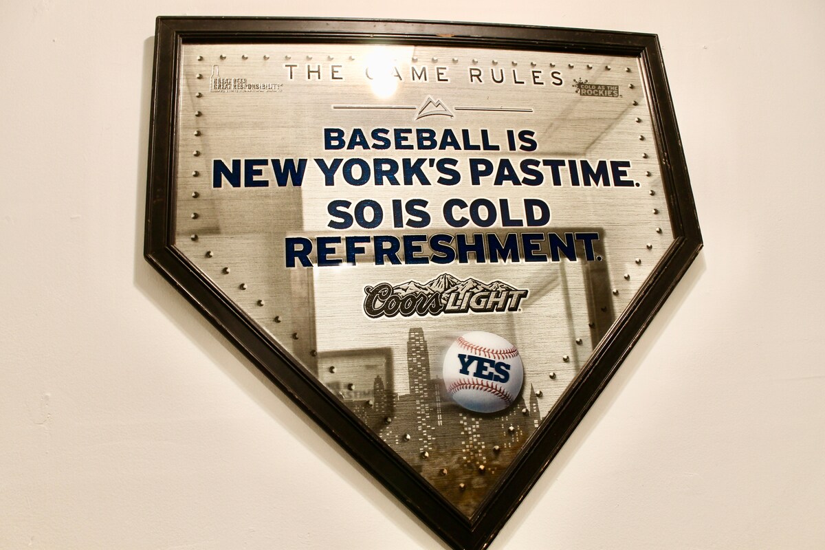 A framed sign in the shape of a baseball home plate, featuring bold text that states 'Baseball is New York's pastime. So is cold refreshment.' The sign is decorated with images related to baseball and beer, reflecting local culture.