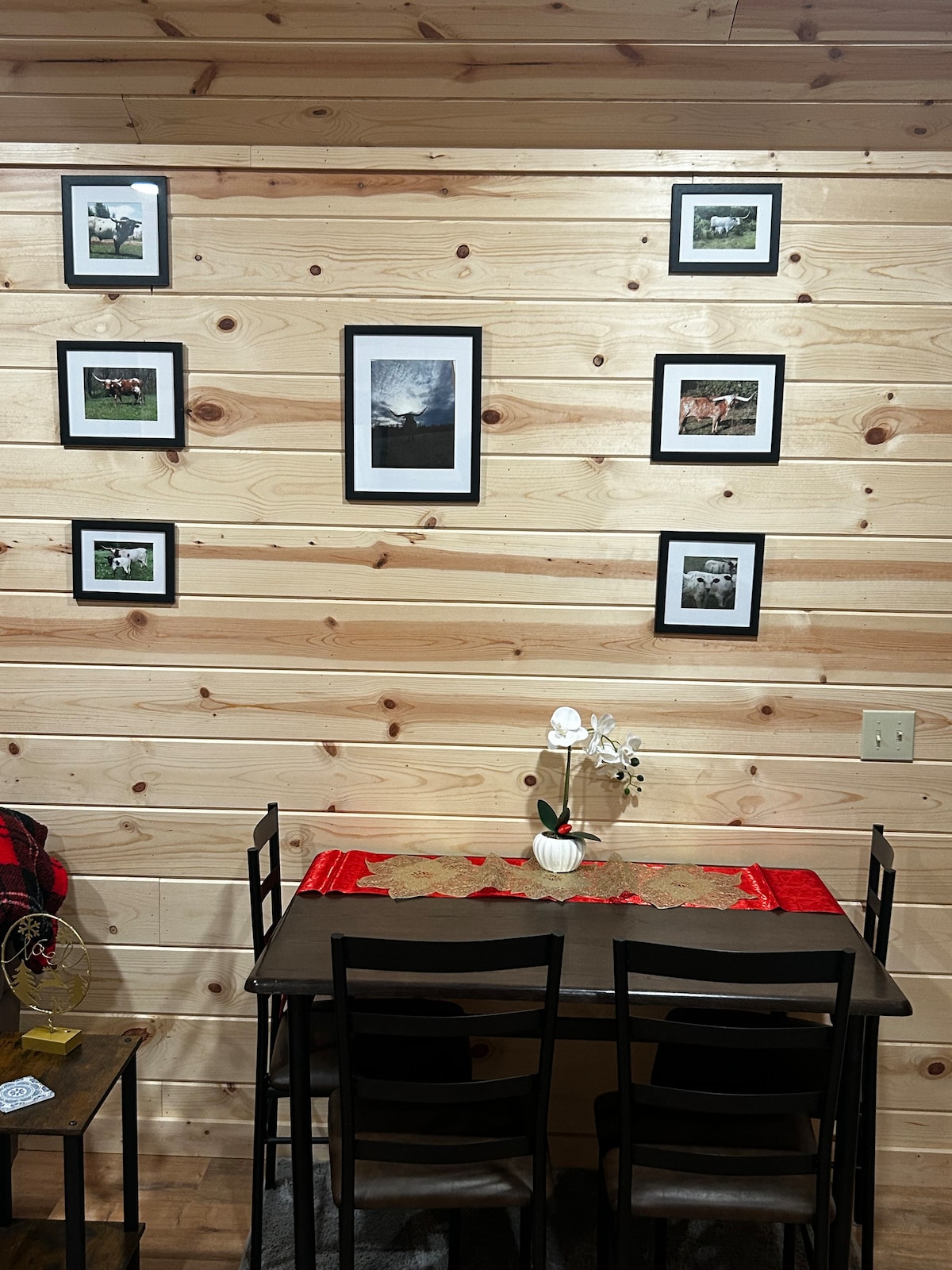 A wooden wall adorned with several framed photographs depicts serene ranch scenes. A simple dining table with four chairs is set against the wall, featuring a decorative table runner and a small flower arrangement as a centerpiece.